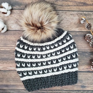 May include: Grey and white knitted beanie with a white pom pom. The beanie has a geometric pattern with white stripes and a grey background.