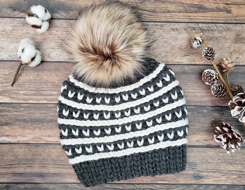May include: Grey and white knitted beanie with a white pom pom. The beanie has a geometric pattern with white stripes and a grey background.