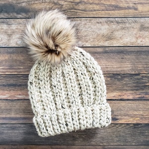 K&ouml;nnte beinhalten: Eine cremefarbene Strickm&uuml;tze mit einem flauschigen braunen Bommel oben. Die M&uuml;tze ist aus einem dicken Garn gefertigt und hat eine gerippte Struktur.