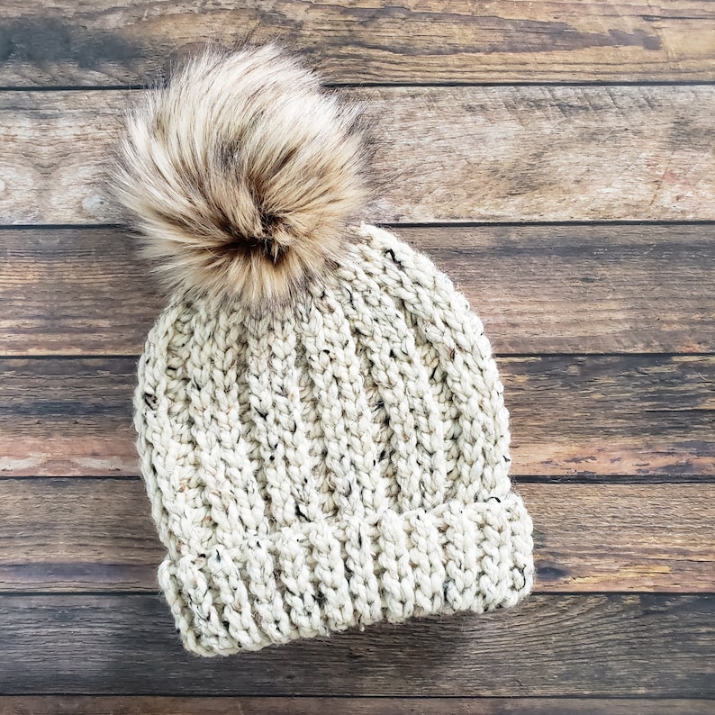 K&ouml;nnte beinhalten: Eine cremefarbene Strickm&uuml;tze mit einem flauschigen braunen Bommel oben. Die M&uuml;tze ist aus einem dicken Garn gefertigt und hat eine gerippte Struktur.