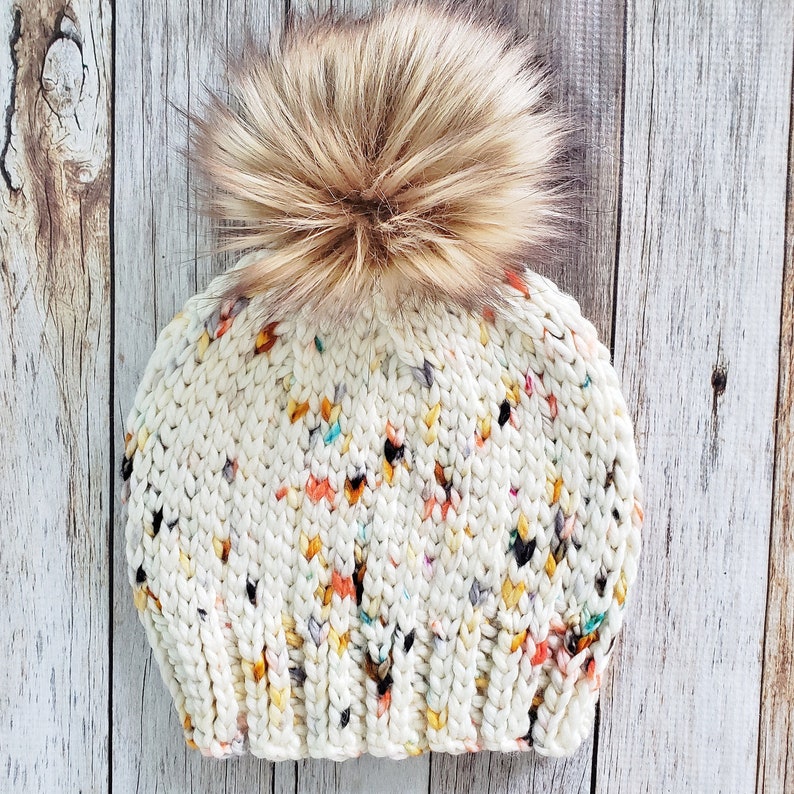 May include: A white knitted beanie with a colourful flecked pattern and a fluffy brown pom pom.