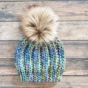 K&ouml;nnte beinhalten: Eine bunte Strickm&uuml;tze mit einem flauschigen braunen Bommel. Die M&uuml;tze ist aus einem dicken Garn gestrickt und hat einen Farbverlauf von Blau, Gr&uuml;n und Lila.