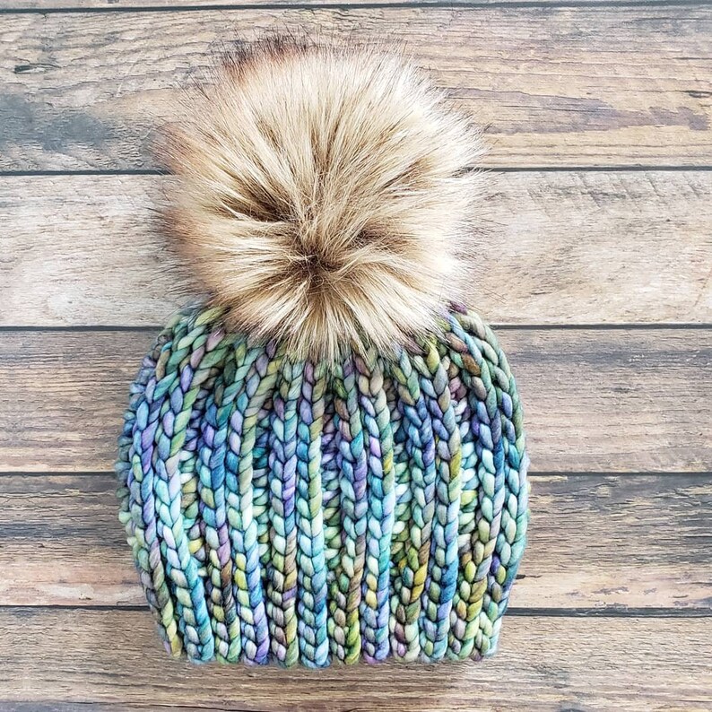 K&ouml;nnte beinhalten: Eine bunte Strickm&uuml;tze mit einem flauschigen braunen Bommel. Die M&uuml;tze ist aus einem dicken Garn gestrickt und hat einen Farbverlauf von Blau, Gr&uuml;n und Lila.