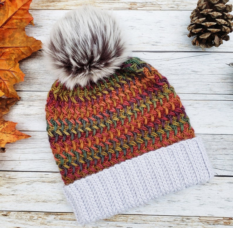 May include: A colorful knit beanie with a faux fur pom-pom. The hat features a woven pattern in shades of red, orange, green, and yellow, with a light gray ribbed brim. The pom-pom is white and brown.
