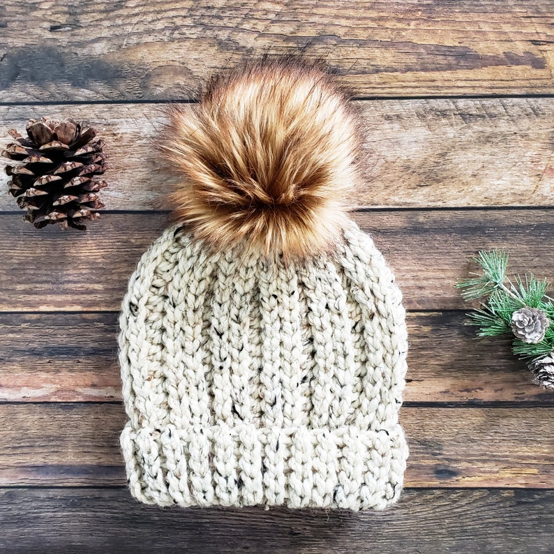 K&ouml;nnte beinhalten: Eine wei&szlig;e H&auml;kelm&uuml;tze mit einem braunen Kunstfellbommel. Die M&uuml;tze hat ein strukturiertes Strickmuster und ist auf einem Holzgrund fotografiert.