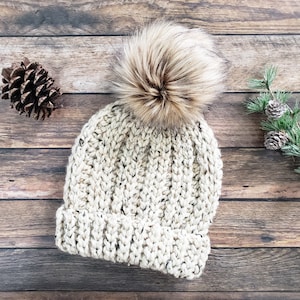 K&ouml;nnte beinhalten: Eine cremefarbene Strickm&uuml;tze mit einem flauschigen braunen Bommel oben. Die M&uuml;tze ist aus einem dicken Garn mit einer gesprenkelten Textur gefertigt.