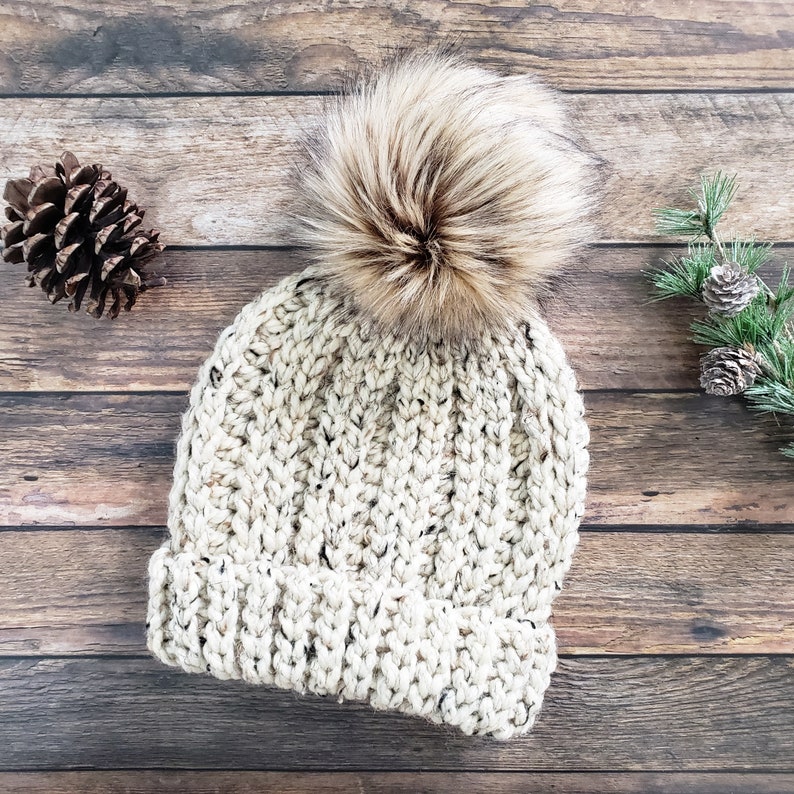 K&ouml;nnte beinhalten: Eine cremefarbene Strickm&uuml;tze mit einem flauschigen braunen Bommel oben. Die M&uuml;tze ist aus einem dicken Garn mit einer gesprenkelten Textur gefertigt.