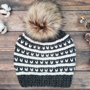 May include: Grey and white striped knitted beanie with a faux fur pom pom. The beanie has a white and grey geometric pattern.
