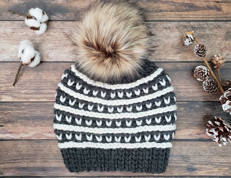 May include: Grey and white striped knitted beanie with a faux fur pom pom. The beanie has a white and grey geometric pattern.