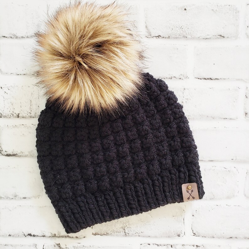 May include: Black beanie with a fluffy brown pom pom and a small brown leather patch with a cross-arrow design.