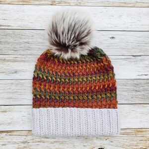 May include: A knitted beanie hat with a faux fur pom-pom. The hat features a textured weave in shades of red, orange, green, and yellow, with a light gray ribbed brim. The pom-pom is a mix of white and brown.
