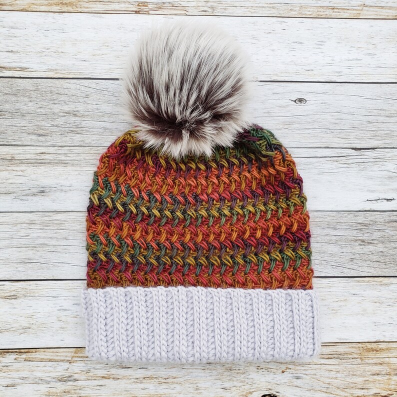 May include: A knitted beanie hat with a faux fur pom-pom. The hat features a textured weave in shades of red, orange, green, and yellow, with a light gray ribbed brim. The pom-pom is a mix of white and brown.