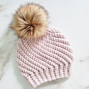 May include: A light pink knitted beanie with a large, fluffy, brown faux fur pom-pom on top. The hat has a ribbed texture and is made of a soft, warm material. The beanie is a winter accessory.