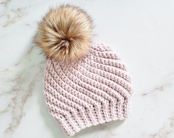 Può includere: Un berretto lavorato a maglia rosa chiaro con un grande e soffice pompon in finta pelliccia marrone. Il cappello ha una trama a coste ed è realizzato in un materiale morbido e caldo. Un accessorio invernale.