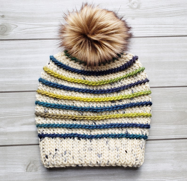 Pode incluir: Um gorro branco de croch&ecirc; com um pompom de pele sint&eacute;tica marrom. O gorro tem listras de fio azul, verde e amarelo.