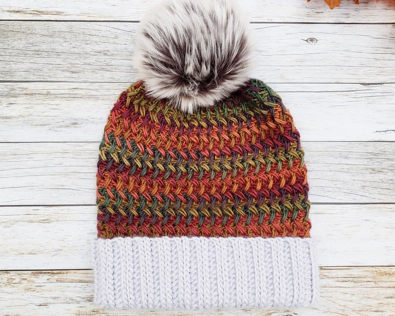 May include: A knitted winter hat with a gray ribbed brim and a colorful woven pattern in shades of orange, green, and red. The hat is topped with a fluffy, faux fur pom-pom in brown and white.
