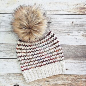 May include: A white crocheted beanie with a multi-colored chevron pattern and a brown faux fur pom pom.