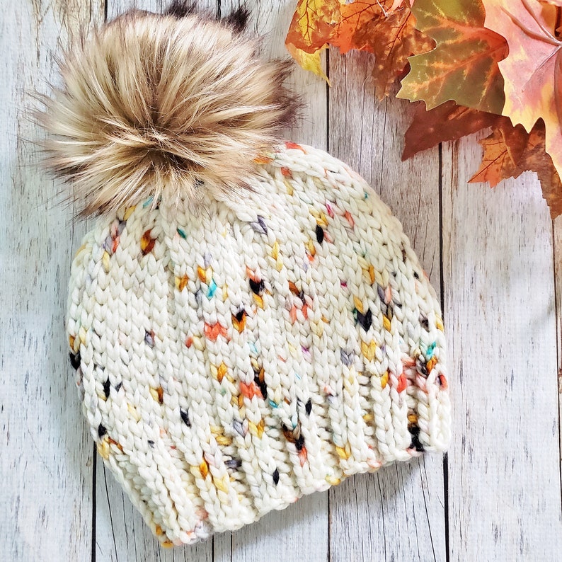 May include: A white knitted beanie with a fluffy brown pom pom. The beanie has a speckled pattern with pops of colour, including yellow, orange, and blue.
