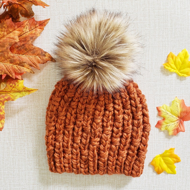 May include: A brown knitted beanie with a fluffy brown pom pom on top. The beanie is made of a thick, chunky yarn and has a ribbed texture.