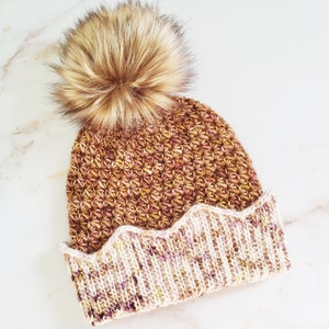 May include: A brown and cream crocheted beanie with a fluffy brown pom pom. The beanie has a scalloped edge.