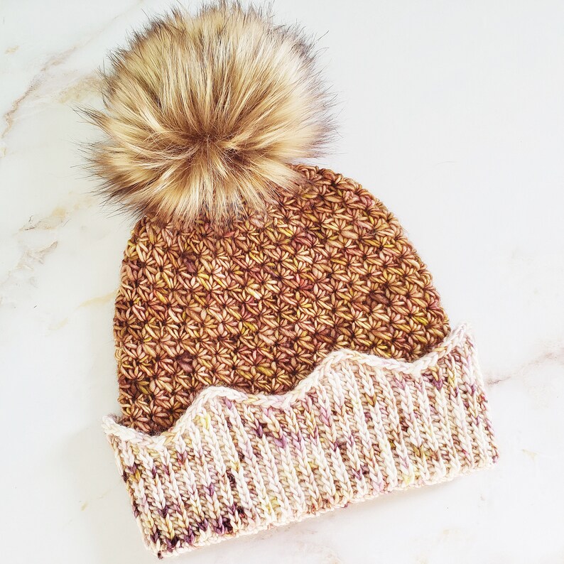 May include: A brown and cream crocheted beanie with a fluffy brown pom pom. The beanie has a scalloped edge.