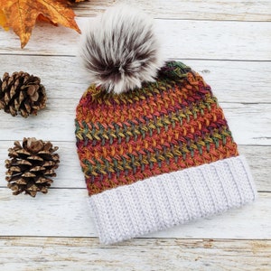 May include: A knitted winter hat with a textured pattern in shades of orange, red, green, and yellow. The hat has a light gray ribbed brim and a fluffy, faux fur pom-pom on top. The hat is displayed on a white wooden surface.
