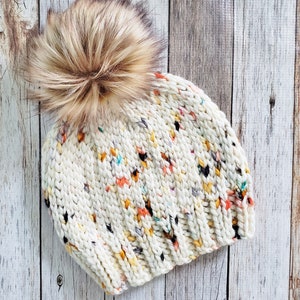 May include: A white knitted beanie with a multi-coloured flecked pattern and a fluffy brown pom pom on top.