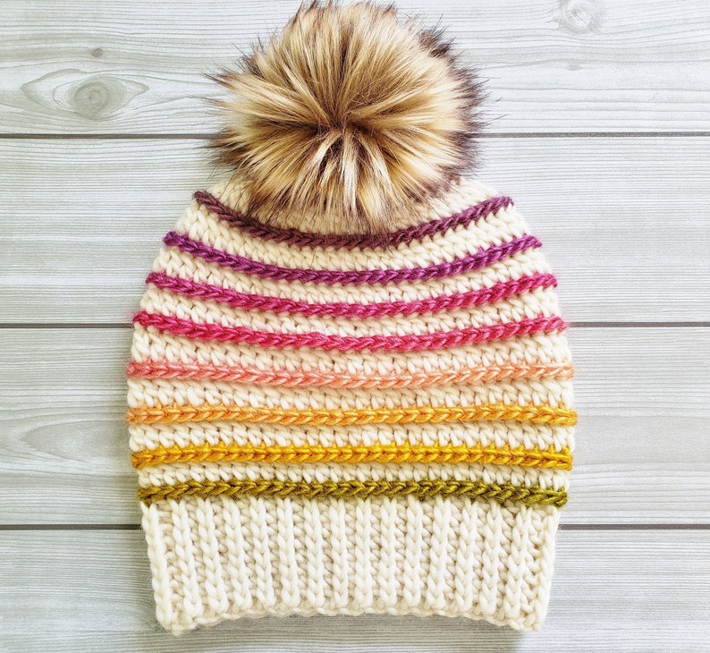 Pode incluir: Um gorro de croch&ecirc; branco com um padr&atilde;o listrado colorido em tons de rosa, roxo, laranja e amarelo. O gorro tem um pompom marrom felpudo no topo.