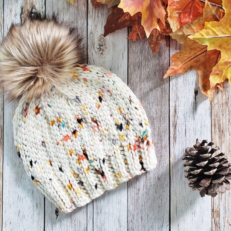 May include: A white knitted beanie with a colourful flecked pattern and a brown faux fur pom pom. The beanie is on a white wood background.