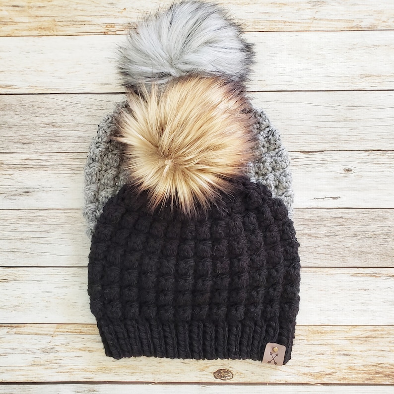 May include: Black crocheted beanie with a gray and brown faux fur pom pom. The beanie has a small brown leather patch with a cross design.