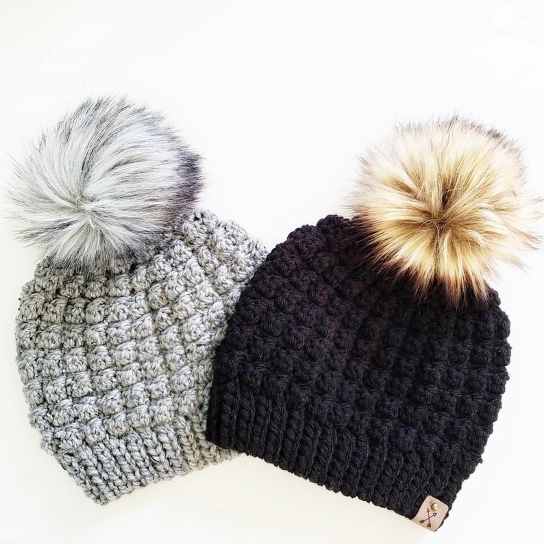 May include: Two crocheted beanies with faux fur pom poms. One beanie is gray and the other is black.