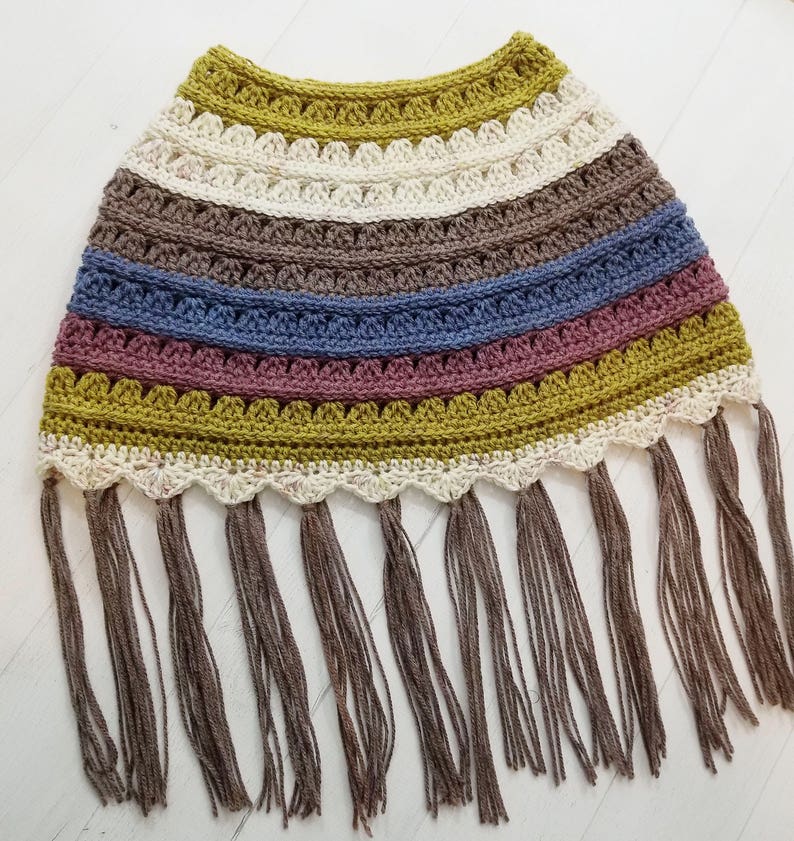 May include: A crocheted poncho with a multi-colored striped pattern in shades of brown, blue, pink, and green. The poncho has a scalloped edge and a fringe trim.