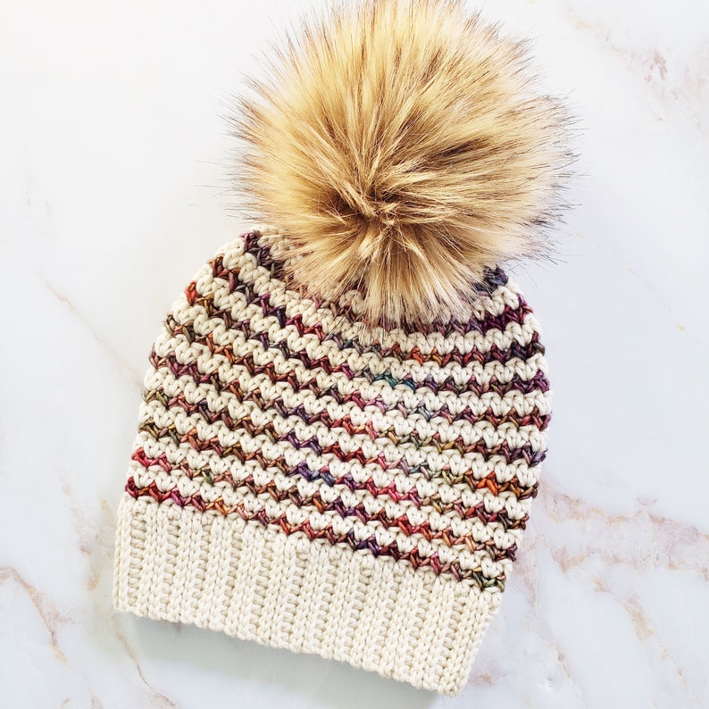 May include: A white crocheted beanie with a multi-colored striped pattern and a brown faux fur pom pom.