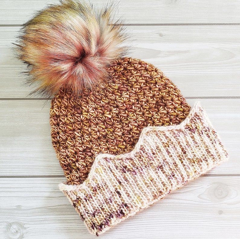 May include: A brown and cream crocheted beanie with a fluffy pink and brown pom pom on top. The beanie has a scalloped edge.