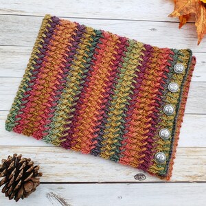 Op de afbeelding: Een kleurrijke gebreide nekwarmer met een textuurpatroon en zilveren knopen. De nekwarmer is gemaakt met verschillende herfstkleuren, waaronder oranje, rood, groen en bruin.