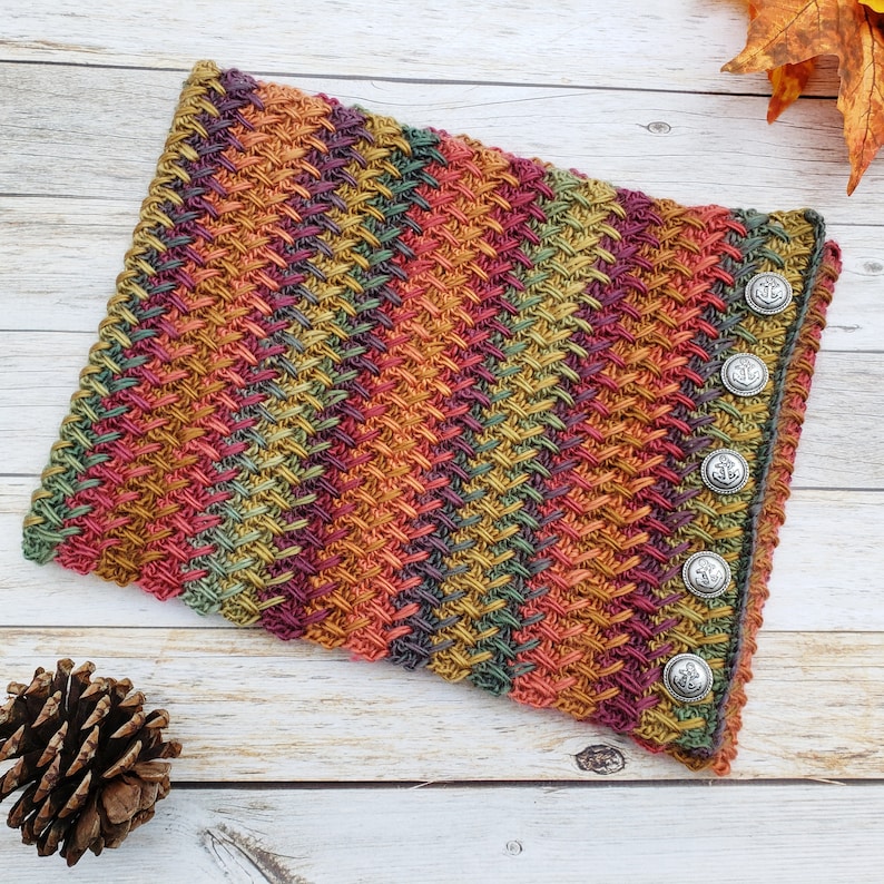Op de afbeelding: Een kleurrijke gebreide nekwarmer met een textuurpatroon en zilveren knopen. De nekwarmer is gemaakt met verschillende herfstkleuren, waaronder oranje, rood, groen en bruin.