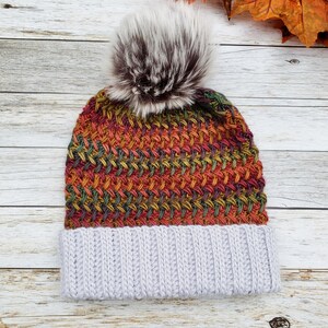 May include: A hand-knitted beanie hat with a textured, colorful pattern in shades of red, orange, green, and yellow. The hat features a light gray ribbed brim and a fluffy, faux fur pom-pom in shades of brown and white.
