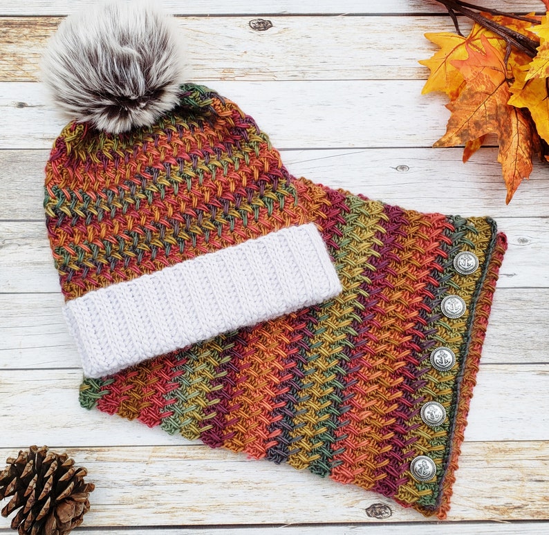 Op de afbeelding: Een gebreide set van muts en sjaal in een veelkleurig patroon van rood, oranje, groen en bruin. De muts heeft een witte pompon bovenop en de sjaal heeft zilveren knopen.