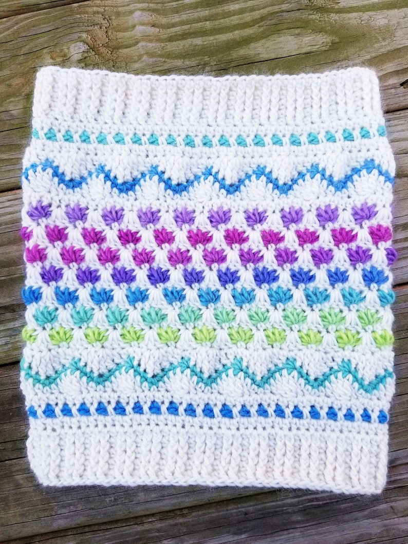 May include: White crochet headband with a colorful chevron pattern and a row of small flower-like stitches in shades of purple, blue, green, and white.