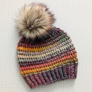 May include: A hand-knitted beanie hat with a large faux fur pom-pom. The hat features a colorful, textured design with shades of pink, yellow, green, and brown. The pom-pom is a mix of light and dark brown.