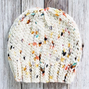 May include: A white knitted beanie with a speckled pattern of orange, yellow, and blue. The beanie is made with a chunky yarn and has a slouchy fit.
