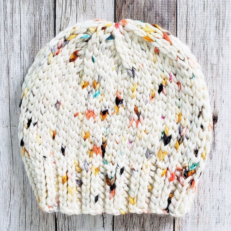 May include: A white knitted beanie with a speckled pattern of orange, yellow, and blue. The beanie is made with a chunky yarn and has a slouchy fit.