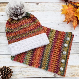 May include: A handmade knit hat and cowl set. The hat has a faux fur pom-pom and a white ribbed brim. The cowl features decorative buttons. The set is in autumn colors of orange, red, green, and yellow.