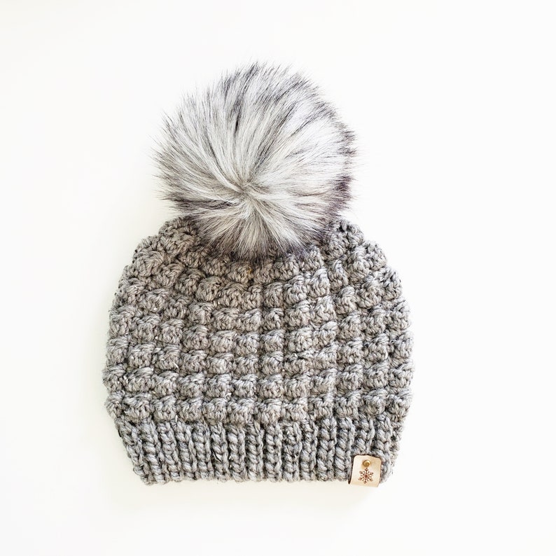 May include: Gray crocheted beanie with a fluffy white pom pom and a small brown leather tag with a snowflake design.