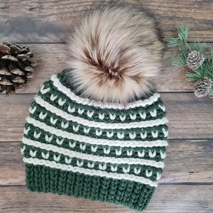May include: A green and white striped crocheted beanie with a faux fur pom pom on top. The beanie is on a wooden background.