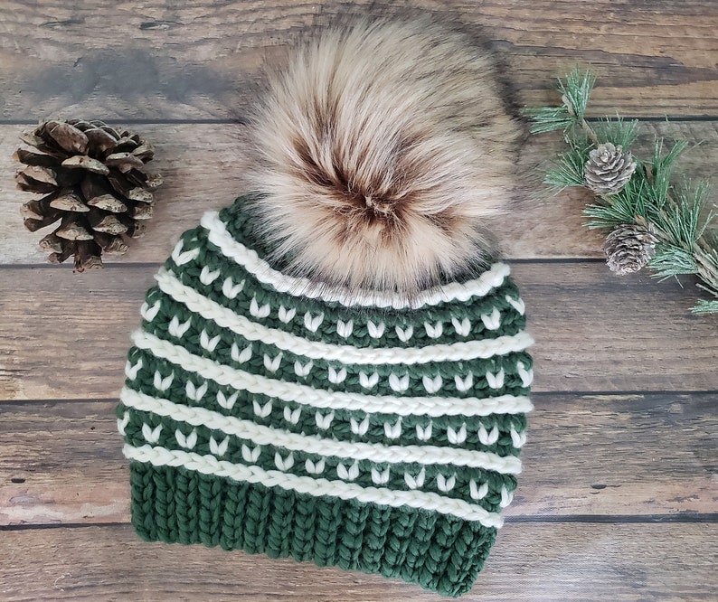 May include: A green and white striped crocheted beanie with a faux fur pom pom on top. The beanie is on a wooden background.