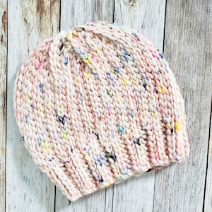 May include: A pink knitted beanie with a speckled pattern of white, yellow, blue, and purple yarn.