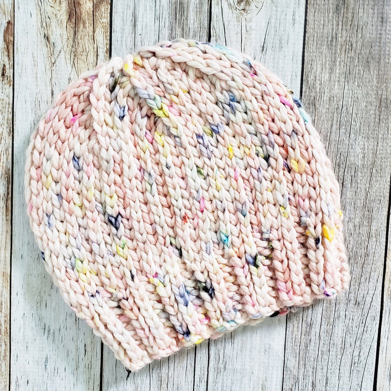 May include: A pink knitted beanie with a speckled pattern of white, yellow, blue, and purple yarn.