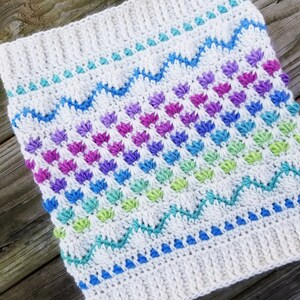 May include: A white crocheted cowl with a rainbow pattern of colorful stitches. The cowl has a textured design with a repeating pattern of small, raised stitches.