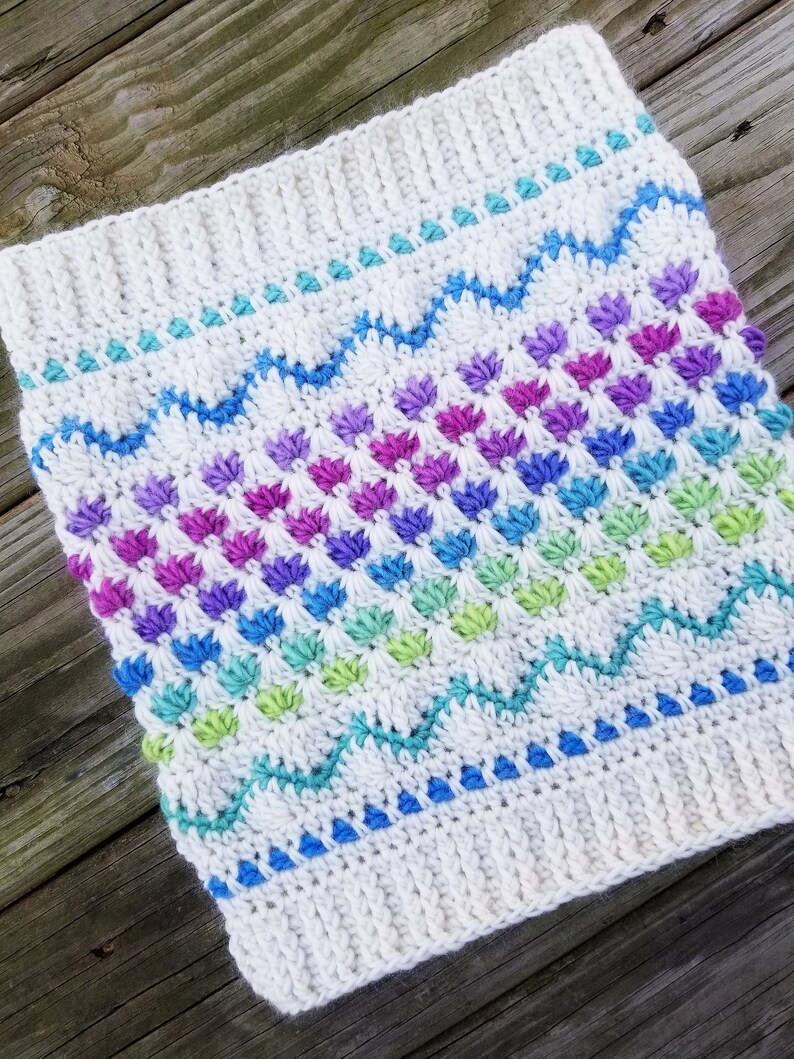 May include: A white crocheted cowl with a rainbow pattern of colorful stitches. The cowl has a textured design with a repeating pattern of small, raised stitches.